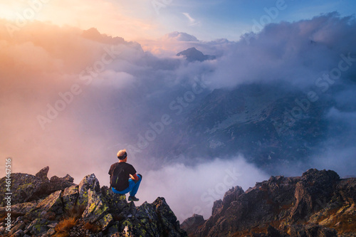 Man sits alone on the peak of rock. Hiker watching to autumn Sun at horizon . Beautiful moment the miracle of nature. Colorful mist in valley. Man hike. Person silhouette stand