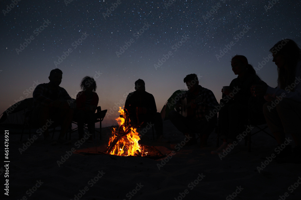Group of friends gathering around bonfire in evening. Camping season - obrazy, fototapety, plakaty 