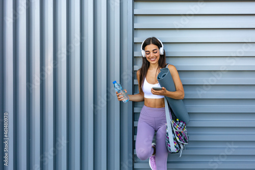 Music and sport. Smiling muscular young woman with headphones  smart watch and bottle of water looks at smartphone on wall background  free space. Athlete runner in sportswear relaxing