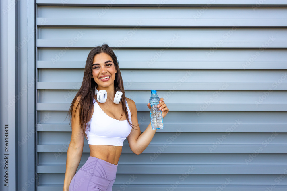 Sporty girl drinking water after workout. Photo of latin fitness girl on grey background. Healthy lifestyle. A beautiful young woman is relaxing during training in summer sunny day drinking the water. Sporty girl drinking water after workout. Photo of latin fitness girl on grey background. Healthy lifestyle. A beautiful young woman is relaxing during training in summer sunny day drinking the water.