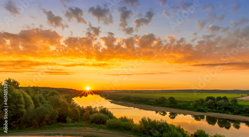Scenic view at beautiful summer river sunset with reflection on water with green bushes, calm water ,deep colorful cloudy sky and glow on horizon on a background, spring evening landscape