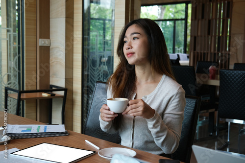 Portrait with beautiful office woman is holding a coffee cup while sitting at the wooden working desk surrounded by a white screen digital tablet and business document.