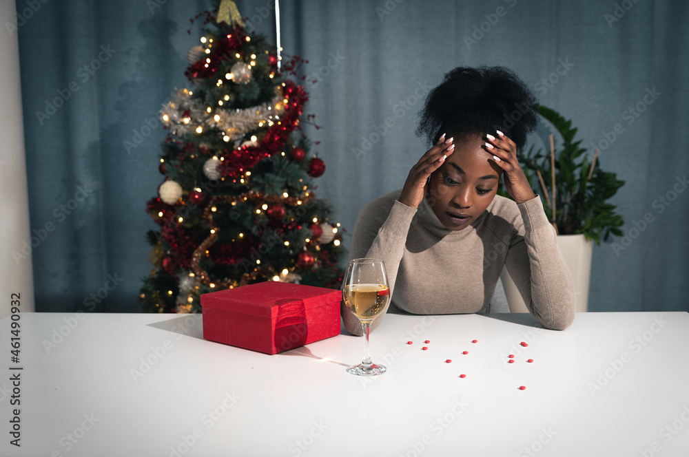 Young unhappy stressed sick young woman sitting at home on Christmas eve with decorated Christmas tree behind drinking champagne and looking at the pain killer pills on the table thinking of a danger Young unhappy stressed sick young woman sitting at home on Christmas eve with decorated Christmas tree behind drinking champagne and looking at the pain killer pills on the table thinking of a danger