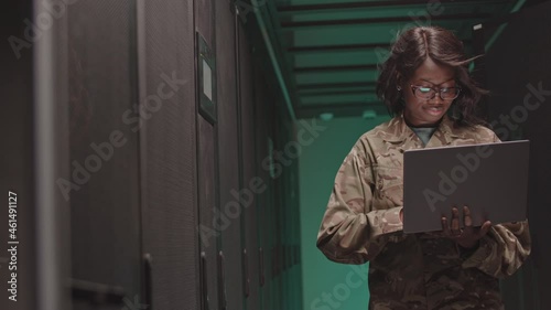 Medium of young African American cyber security specialist holding laptop computer, typing, standing in green-lighted server room, cooling fan blowing on her hair photo