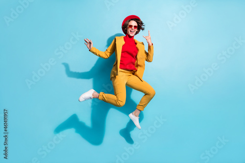 Photo of pretty cool young lady wear yellow blazer red cap smiling jumping showing hard rock sign isolated blue color background
