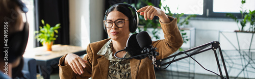 asian radio host in glasses and headphones gesturing near blurred colleague in studio, banner