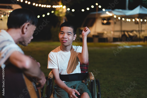A person with disability in wheelchair singing and father playing guitar happily at the camp night, Lifestyle of happy teen vacation with family.