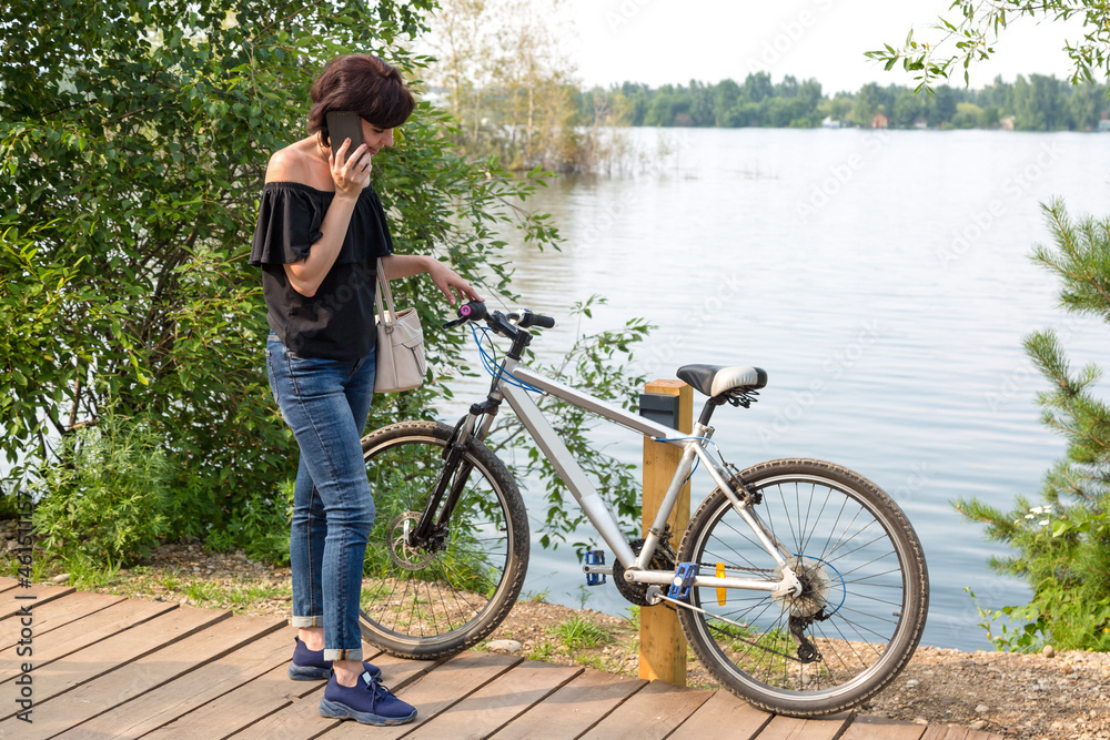 Fototapeta premium frustrated woman with broken bicycle stands by the river and calls her husband waiting for help.
