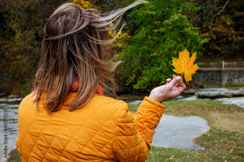 Woman in yellow jacket keep yellow orange maple leaf. Hair tossed lightly in the breeze. Forest background