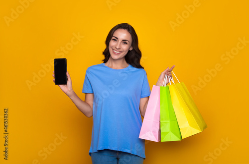 Happy smiling young woman in casual clothes with colorful shopping bags is showing smart phone and posing on yellow background