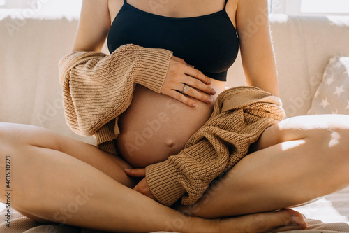 Closeup of a pregnant woman sitting on a sofa in soft light, hugging her belly, wearing a cozy cardigan.