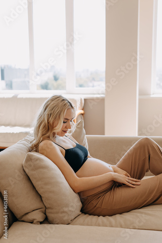 Pregnant woman wearing cozy home wear, lying on a sofa in a light interior at home.