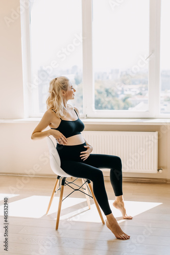 Blonde pregnant woman wearing black top and leggins, sitting on a chair at home, looking through the window.