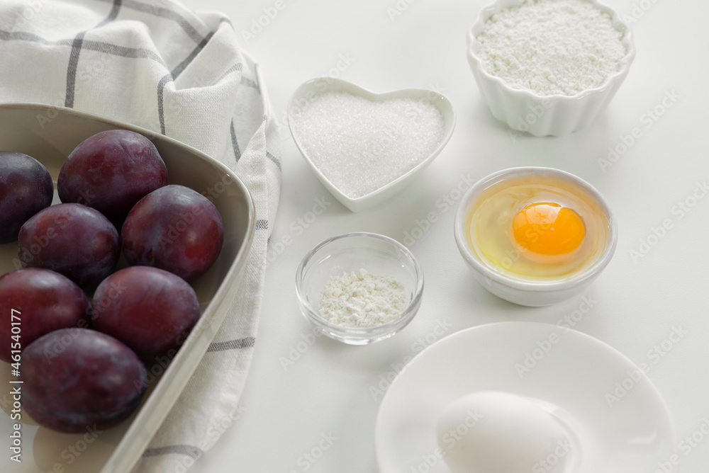 Fototapeta premium Step 1: flour, eggs, sugar, plums for sweet pastries on white background. Step by step recipe for baking sweet plum pie. Ingredients set for cooking. Culinary light background