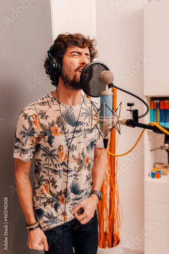 Young caucasian musician singing into microphone while working on songs during a recording studio session at home. Grey background. Millennial singer using headphones  recording his voice.