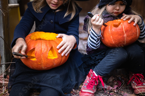 Little girls make jack-o-lantern from big pumpkins for celebratiion of halloween holiday.Witch costume, hat, coat. Cut with knife,take out pulp with seeds.Outdoors activity, backyard.Children's party photo