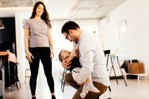 Young father holding the son in his arms and while pregnant mother watching them in their home