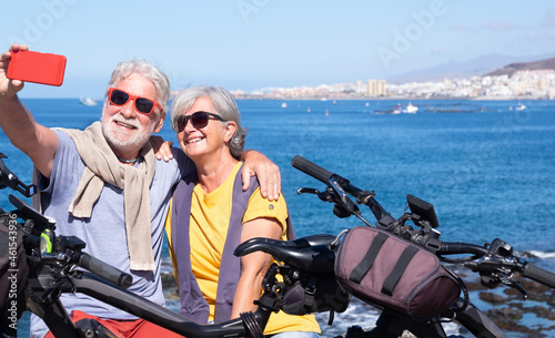 Smiling beautiful elderly couple sitting on the cliff taking a selfie with smartphone. Active retirees enjoying healthy lifestyle and freedom using bicycles - horizon over water