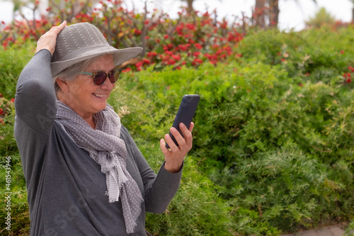 Portrait of beautiful senior woman with hat standing in public park using mobile phone in video call. Smiling elderly lady gray dressed . Bushes and flowers in background