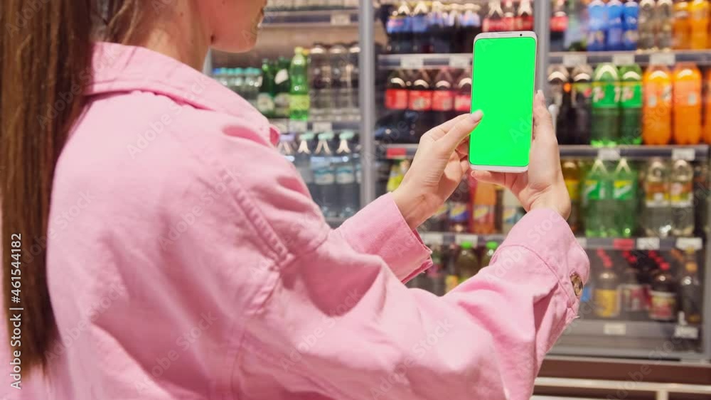 Young woman holding mobile phone in her hands making photo while standing in sparkling water or lemonade store near the fridge.