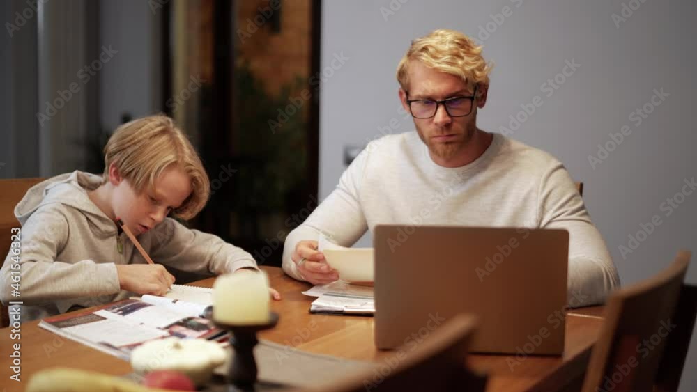 Serious father working with papers while his son do homework at home Serious father working with papers while his son do homework at home