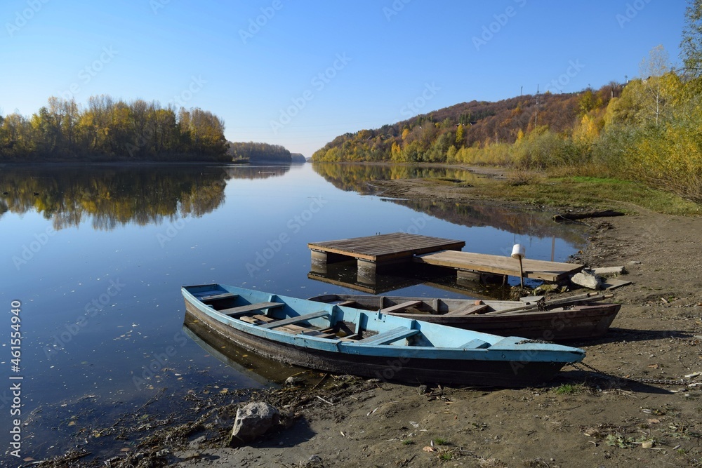 On the river bank near the pier there are two wooden boats against the background of the autumn forest
