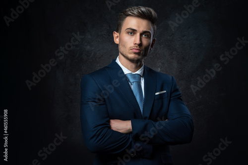 Conceptual portrait of a young, stylish business man, in a blue suit, hands on a cross, against a dark textured background.