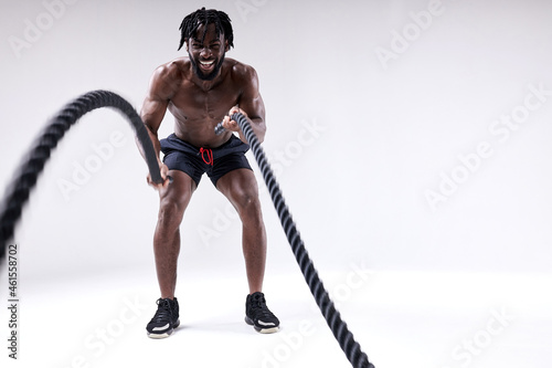 Athlete young black man doing cross fit exercises with battle ropes over isolated white background. hard workout of african shirtless guy. full-length portrait. sport and motivation concept