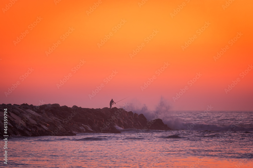 Fisherman silhouette fishing in the atlantic ocean with waves crashing on a stone pier at sunset, in Portugal