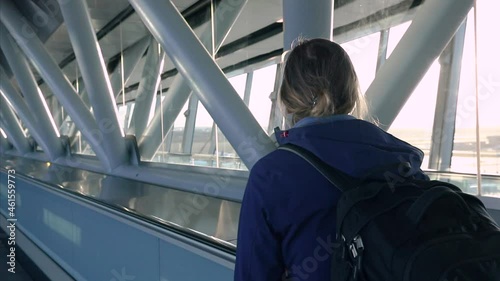 Passenger on airport moving walkway going to flight boarding gate. Young woman traveler with backpack. International terminal interior with sunset light.