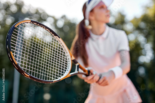 Pretty redhead female is playing tennis, woman is waiting for the serve, looking at side, wearing white uniform and cap, side view portrait. Slim athlete lady is engaged in sport, focus on racket