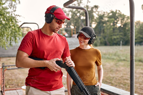 Confident male shooting instructor teaches the backslider how to handle weapon on training course in outdoor range. positive teacher and woman in protective spectacles and headset have fun, laugh