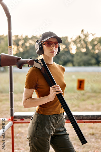 Beautiful caucasian lady with firing gun in outdoor academy shooting range, field in the background. Young beautiful woman in cap, headset and spectacles posing at camera after successful training