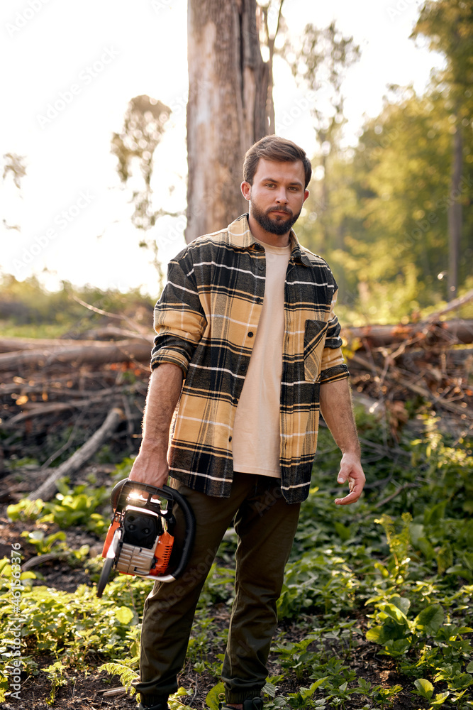 Serious young caucasian lumberjack in casual shirt is preparing to cut  trees with electric saw, standing in forest alone, posing, at summer  evening. outdoor activity. strong man with saw. woodcutter. foto de