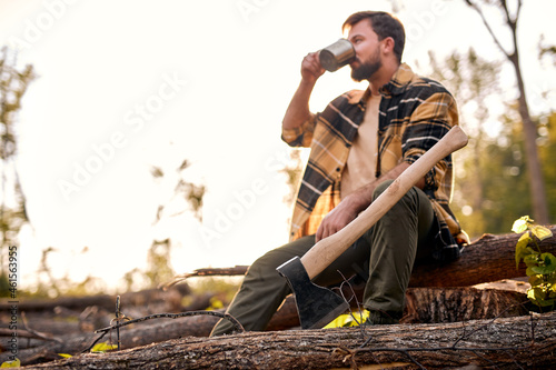Detail of axe. Axe and firewood. Sharp ax standing on a wooden, young caucasian man in casual shirt in the background. handsome tired guy sit drinking hot tea, having rest after chopping