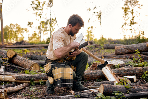 Side view on young handsome lumberman collecting prepared chopped firewood in forest, Wood worker in casual outfit engaged in forestry at summer season alone. Wood work, handwork, timber