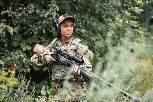 confident woman in camouflage clothing and bulletproof vest stands in field against the background of forest. young military woman holds an automatic rifle in hands, posing, alone. outdoor portrait