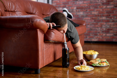 Fat Lazy Boy Taking Fries From Plate Lying On Sofa, Alone At Home, Teenager Boy Having Rest After School, Relaxed. Caucasian child lead unhealthy lifestyle, eating junk meal. overweight, obesity photo