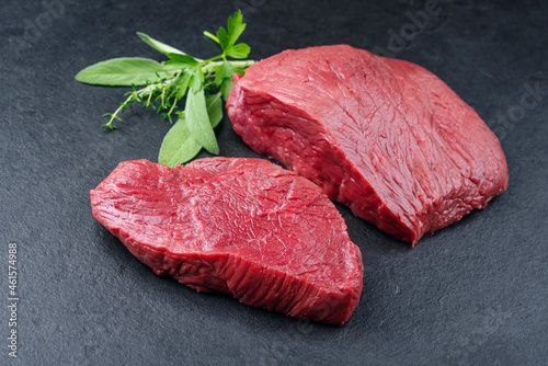 Raw dry aged bison beef rump steak piece and slices with herbs offered as close-up on black background photo