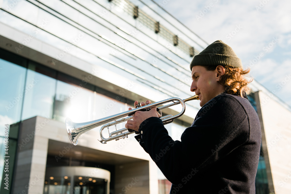 Obraz premium A man with a hat playing a trumpet on modern city background. Male Jazz musician play music on city streets and looking to the side 