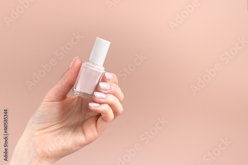 Female hand holding a pink nail polish bottle on a soft beige background. Natural manicure presentation. Luxury woman's cosmetics concept