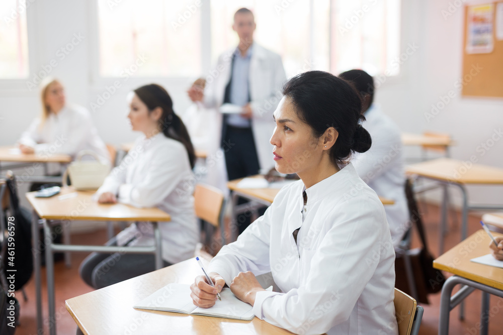 Young positive woman in white medical coat sitting at desk in classroom attending seminar