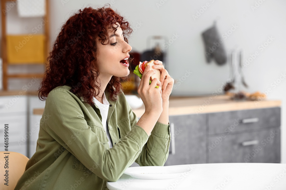 Pretty young woman eating tasty sandwich at table in kitchen Pretty young woman eating tasty sandwich at table in kitchen