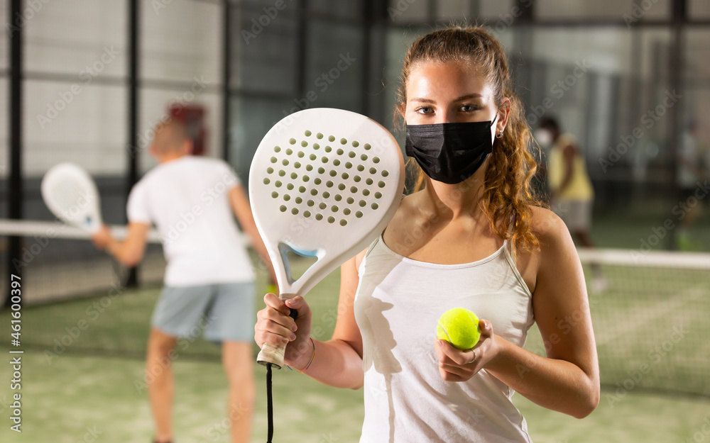 Portrait of confident sporty girl wearing protective mask standing on paddle tennis indoor court with racket and ball in hands. Precautions in coronavirus pandemic