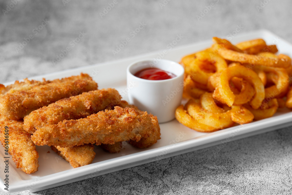 Chicken goujons, coated in breadcrumbs, with spicy curly fries and a tomato ketchup dip on a white platter. On a concrete background Chicken goujons, coated in breadcrumbs, with spicy curly fries and a tomato ketchup dip on a white platter. On a concrete background