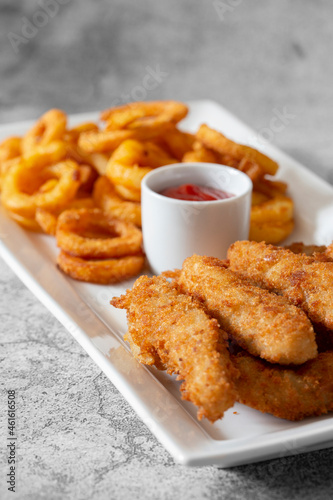 Chicken goujons, coated in breadcrumbs, with spicy curly fries and a tomato ketchup dip on a white platter.  On a concrete background photo