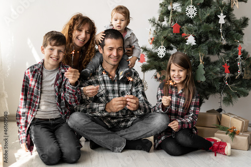 Happy full family and three children at home with Christmas gifts sitting at home on the floor in the morning under a Christmas tree in winter.