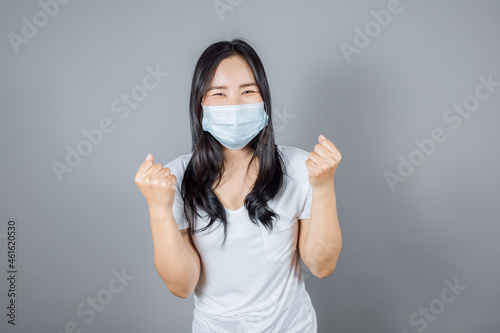 Portrait of happy Asian woman wears white t-shirt in medical face mask to protect Covid-19  Coronavirus  raise hands glad excited cheerful on gray background