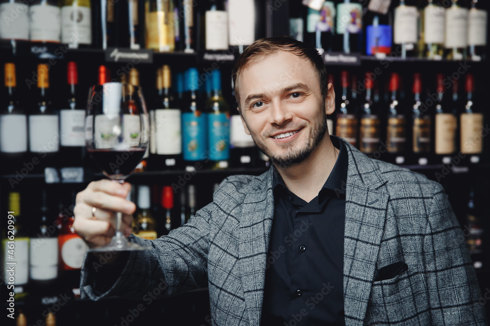 Portrait of happy man with glass of red wine on background of showcase of alcohol store