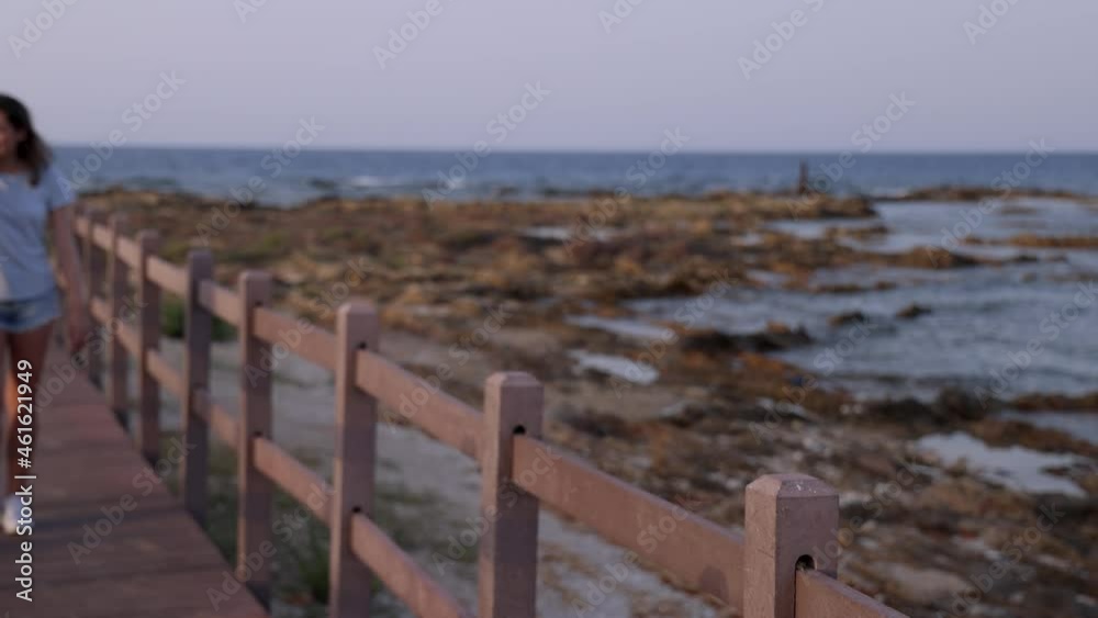 Mother and daughter walking up the wooden pier by the sea at sunset and talking. A middle-aged woman and teenage girl wearing blue t-shirts and shorts. Motherhood concept, closeness to nature concept Mother and daughter walking up the wooden pier by the sea at sunset and talking. A middle-aged woman and teenage girl wearing blue t-shirts and shorts. Motherhood concept, closeness to nature concept
